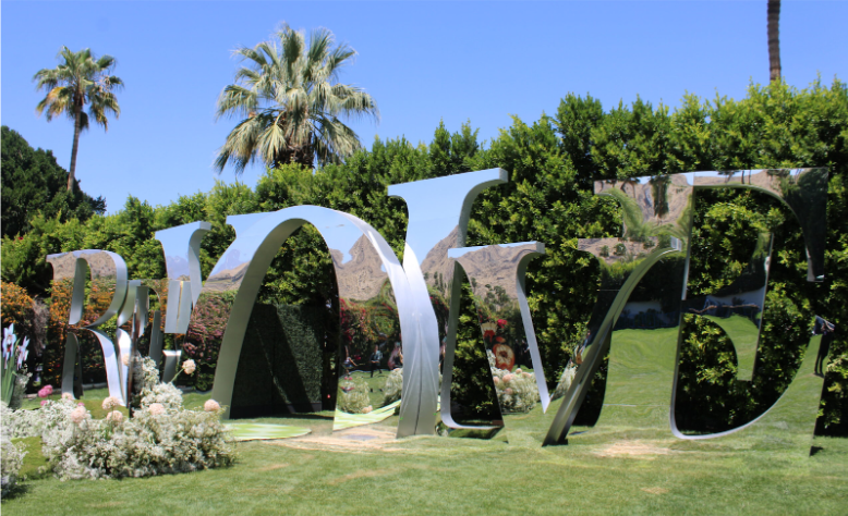 This stunning mirrored installation spelling out ‘Revolve’ stands amidst a lush garden, reflecting the vibrant greenery, palm trees, and a clear blue sky at the Revolve Festival. 