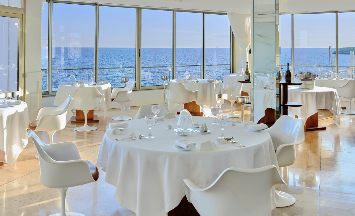 Dine amidst a pristine white ambiance at Le Petit Nice Alt Text: Embrace Le Petit Nice's bright, modern interior with pristine white tables and chairs and large windows overlooking a vast ocean view under a clear blue sky.