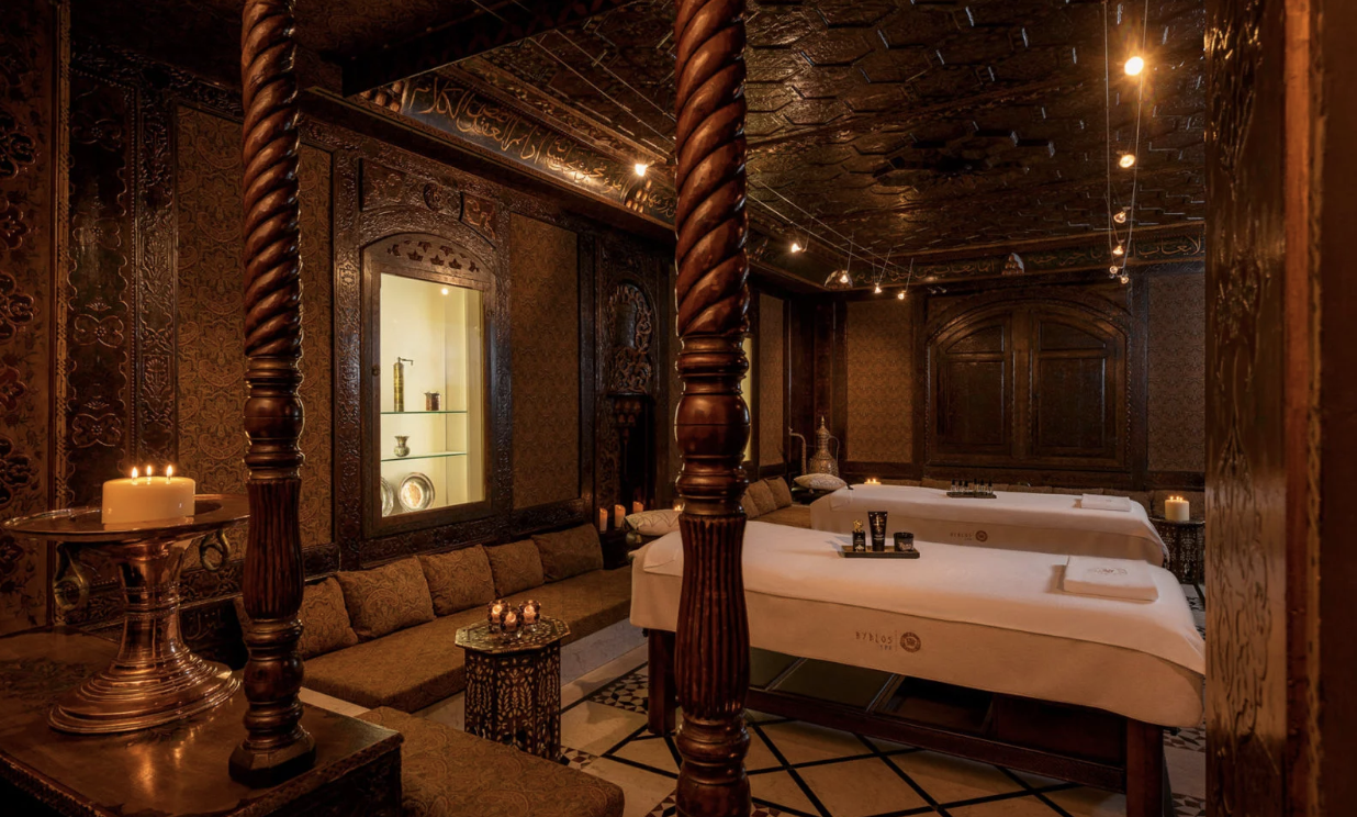 Experience the revival of beauty, body, and mind at Byblos Spa Alt Text: Byblos Spa’s luxurious spa room with ornate wooden decor, dim lighting, candles, and two white linen massage tables, ideal for a peaceful day of relaxation.