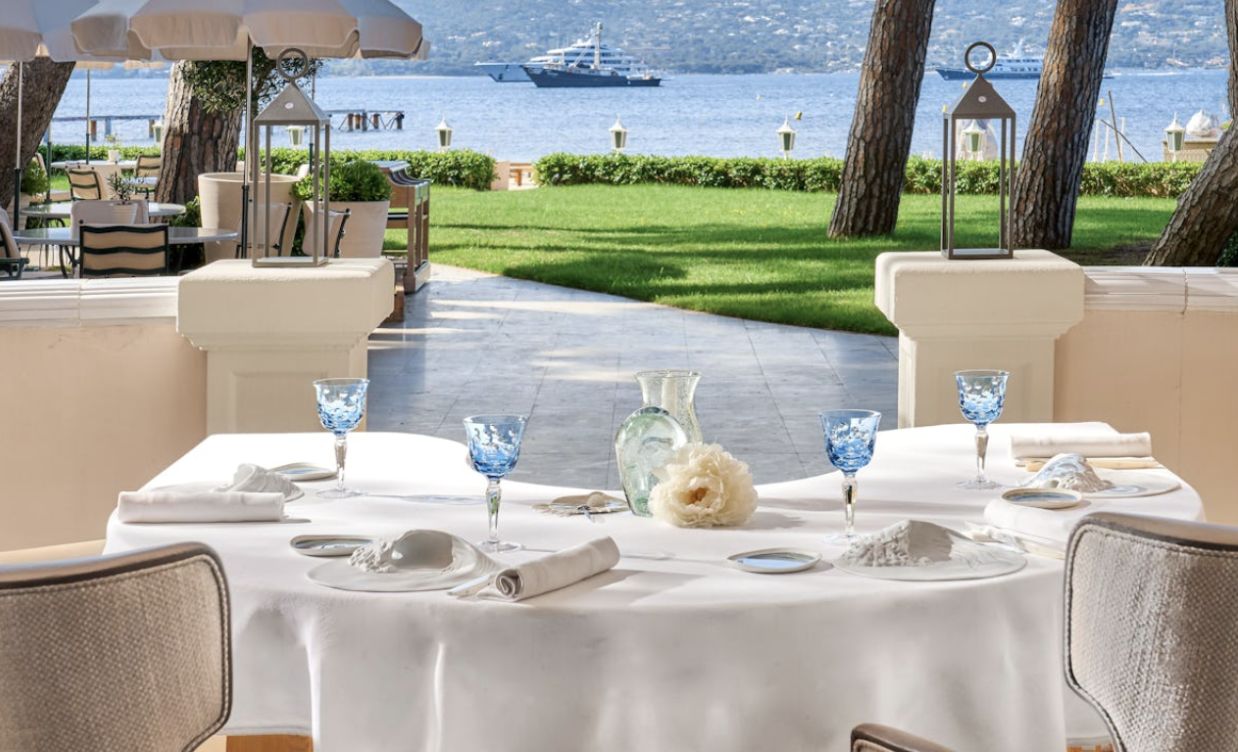 Enchanting view from the terrace at La Vague d’Or Alt Text: A table at La Vague d’Or set with turquoise glasses and contemporary plates overlooking a garden and tranquil water.