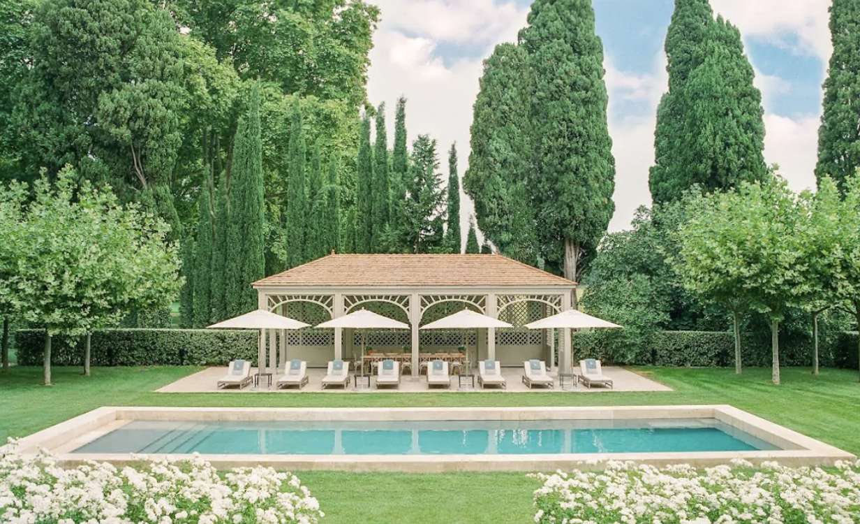 A serene garden at Chateau Avignon Alt Text: Chateau Avignon’s tranquil swimming pool nestled among lush trees and manicured lawns dotted with sunbeds and umbrellas with a pergola as its focal point.