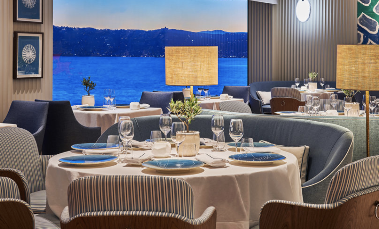 Caption: Breathtaking views from the elegant LouRoc Restaurant  Alt text: A refined dining space overlooking the Mediterranean at sunset, where soft lighting, plush seating, and elegantly set tables create an ambiance of understated luxury. 