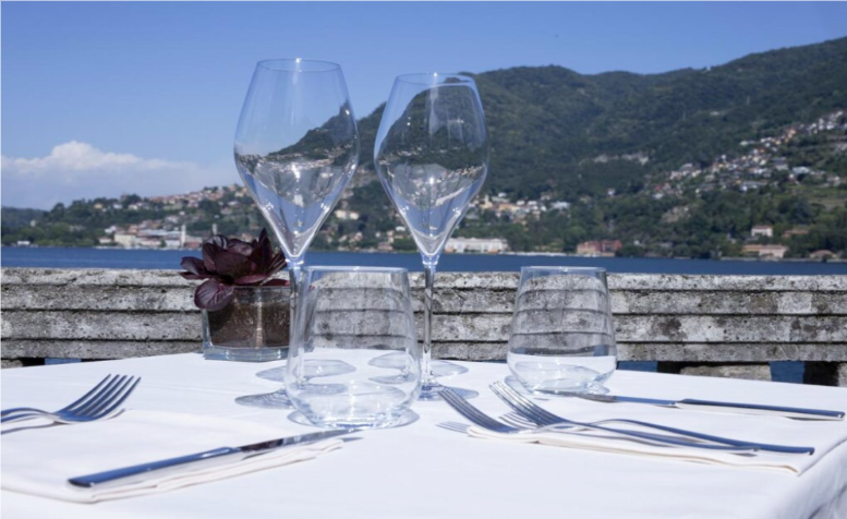 Elegant lakeside dining at Momi restaurant on Lake Como, featuring a beautifully set table with glassware and silverware overlooking the water and surrounding hills.