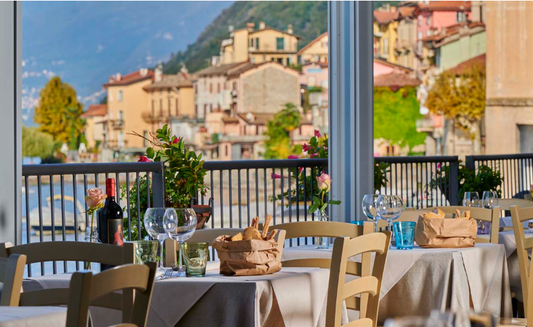 Lakeside dining at Aurora Lezzeno restaurant on Lake Como, featuring an open-air terrace with elegantly set tables overlooking the water and mountains at sunset.