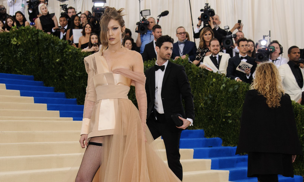 Gigi Hadid poses on the 2017 Met Gala red carpet in a dramatic nude-toned couture gown with a flowing tulle train, surrounded by photographers.