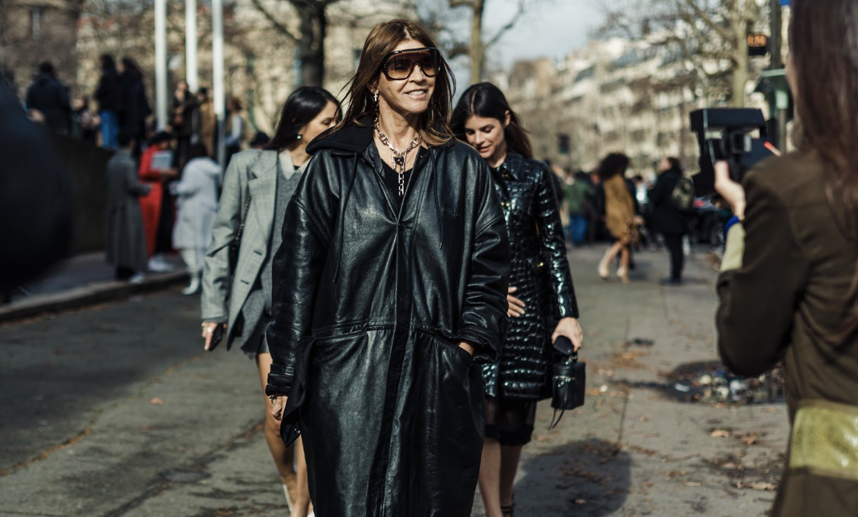 Walking confidently down a lively city street, Carine Roitfeld wears an oversized black leather coat and sunglasses.