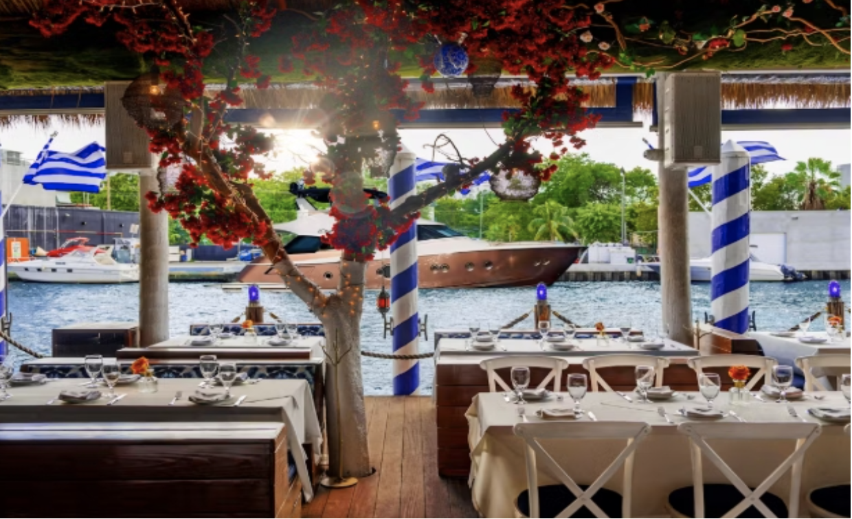 Outdoor waterfront seating at Kiki on the River in Miami, featuring Greek-inspired decor, dockside dining, and yachts moored nearby.